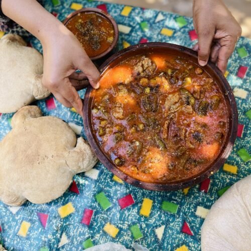 A clay pot of stew with flatbread on a patterned cloth at a farm table