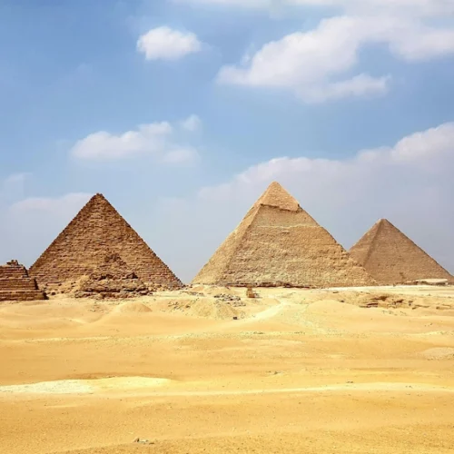 The three pyramids of Giza rising from the sand under a blue sky with scattered cloud