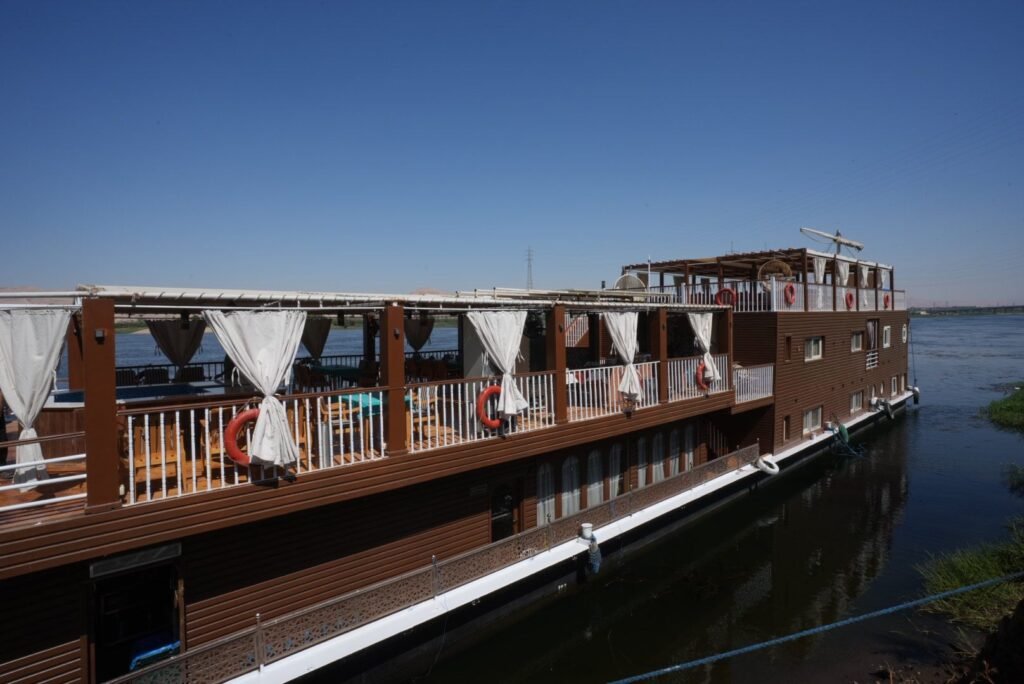 A two-deck wooden dahabeya moored on the Nile, curtained upper deck and the lateen yard stowed above the stern