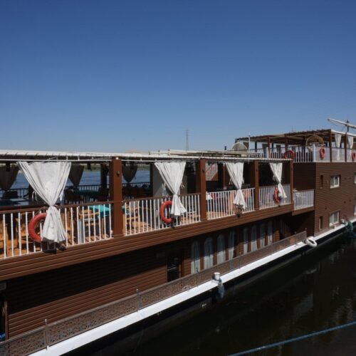 A two-deck wooden dahabeya moored on the Nile, curtained upper deck and the lateen yard stowed above the stern