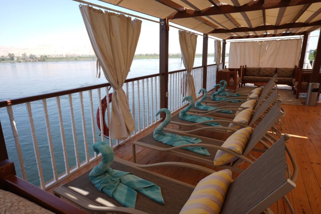 Sun loungers under a wooden pergola on the upper deck of a Nile dahabeya, the river and the west bank beyond